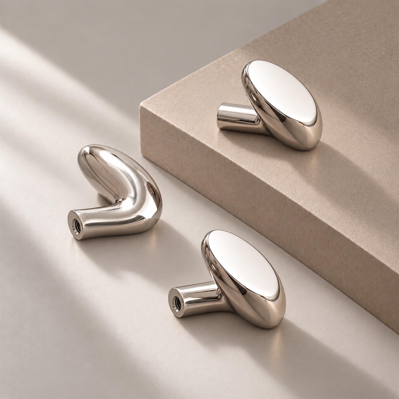 May include: Three polished silver-toned metal cabinet knobs with a smooth, rounded, and organic shape. Each knob features a cylindrical base with a threaded hole for mounting, showcasing a reflective, high-gloss finish and a curved, pill-like head.
