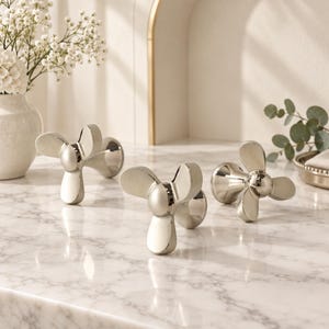 May include: Three polished chrome faucet handles, each featuring a distinctive three-blade propeller design. These metallic fixtures have a smooth, reflective finish and are mounted on circular bases, showcasing a modern and functional aesthetic.