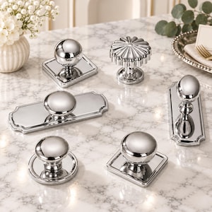 May include: A collection of six polished chrome cabinet knobs and pulls with reflective surfaces. The set includes various shapes such as round, square, and rectangular backplates, along with one fluted circular knob, all featuring a bright metallic finish.