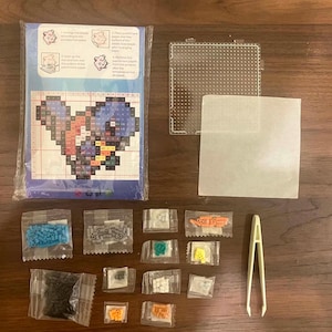 May include: A fuse bead craft kit including a blue instruction sheet with a pixelated character pattern, a clear square pegboard, a sheet of parchment paper, multiple small plastic bags containing assorted colored beads, and white plastic tweezers.