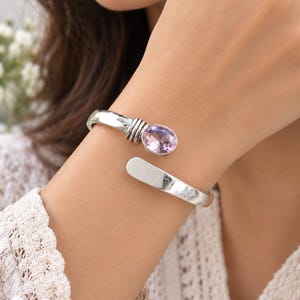May include: A silver-toned open cuff bracelet featuring a polished, smooth band with a ribbed metal detail near one end. The bracelet is accented by a single, oval-shaped, faceted light purple gemstone set in a silver-toned metal bezel setting.