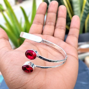 Pode incluir: Uma pulseira de punho aberto em tom prateado com uma textura de metal martelado e duas pedras vermelhas ovais facetadas em cada extremidade. A pulseira tem um design cruzado com uma secção retangular plana e lisa no topo da banda.
