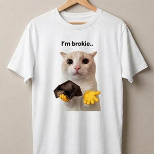 Könnte beinhalten: Ein weißes Kurzarm-T-Shirt mit einer Grafik einer cremefarbenen Katze mit großen Augen, die eine leere dunkelbraune Brieftasche hält. Die Katze hat gelbe Cartoon-Hände und der Text "I'm brokie.." ist in Schwarz über dem Kopf der Katze gedruckt.