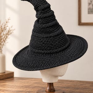 Pode incluir: Um chapéu pontiagudo preto feito com um padrão de crochê denso. O chapéu apresenta uma aba larga e circular e uma coroa cónica alta e ligeiramente curva que termina numa ponta arredondada, exibindo uma textura uniforme em toda a peça.