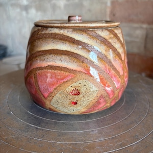 May include: A round ceramic jar with a lid, featuring a speckled beige base with swirling bands of brown and pink glaze. The front displays a small red stamped lotus flower icon above a tiny red heart shape, all finished with a glossy texture.