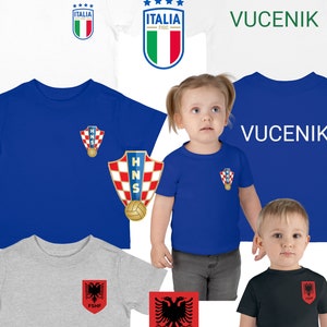 Puede incluir: Cinco camisetas con diferentes escudos de países y nombres de equipos. Las camisetas son de varios colores, incluyendo blanco, azul, gris y negro. Los escudos incluyen la bandera italiana, el escudo del equipo nacional croata y el escudo del equipo nacional albanés. Los nombres de los equipos son "ITALIA FIGC", "VUČENIK", "HNS" y "FSHF".