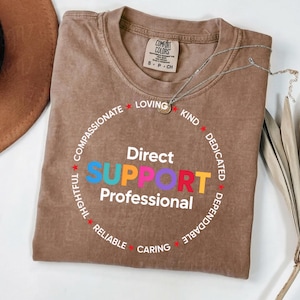 May include: A brown short-sleeved t-shirt with a crew neckline. The front features the white text Direct Support Professional and the word SUPPORT in blue, orange, pink, purple, red, and yellow. A circular border of white text includes words like Loving, Kind, Dedicated, Dependable, Caring, Reliable, Thoughtful, and Compassionate.