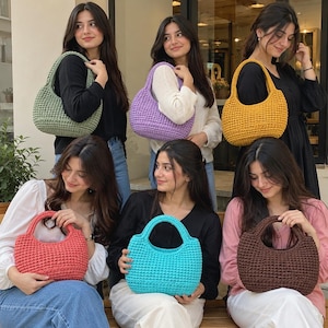 May include: A collection of six crescent-shaped crochet handbags in various solid colors including sage green, lavender, mustard yellow, coral pink, bright turquoise, and dark brown. Each bag features a single integrated handle and textured stitch.