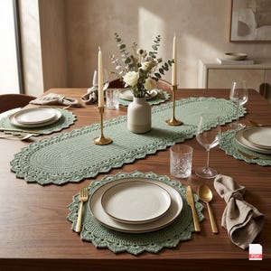 Könnte beinhalten: Ein salbeigrüner gehäkelter Tischläufer und passende runde Platzsets mit gewelltem Rand. Das Set ist auf einem Holztisch mit weißen Keramiktellern, goldfarbenem Besteck, klaren Gläsern und zwei hohen Kerzen in Messinghaltern arrangiert.