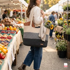 Puede incluir: Un bolso tote grande y rectangular tejido a mano en crochet, con un diseño de bloques de color horizontales en crema, gris claro y gris carbón oscuro. El bolso tiene dos correas gruesas para el hombro, una etiqueta de marca y girasoles.