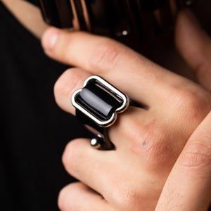 Könnte beinhalten: Ein Ring mit einem glatten, schwarzen, rechteckigen Mittelstück, das von einem polierten, silberfarbenen Metallrand umrahmt wird, mit einem kleinen silberfarbenen Kugelakzent an der Seite der Ringschiene, passend zu einem schwarzen Halsband.