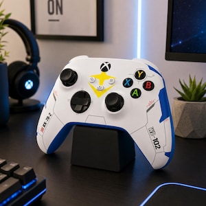 Può includere: Un controller wireless per videogiochi bianco con accenti blu, un emblema a stella gialla e joystick neri. Il controller è contrassegnato con il testo RX-78-2 e WB-102 ed è posizionato su un supporto nero, mostrando i pulsanti standard.