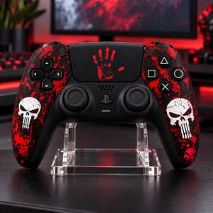 Può includere: Un controller per videogiochi wireless nero con un motivo a schizzi di vernice rossa, grafiche di teschi bianchi sulle impugnature e un'icona di impronta di mano rossa sul touchpad centrale, dotato di pulsanti direzionali e levette analogiche.