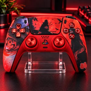 Puede incluir: Un mando de videojuegos inalámbrico de color rojo con un estampado gráfico personalizado que muestra una figura silueteada, pájaros volando y un diseño de ojo grande. Incluye joysticks analógicos rojos metálicos, cruceta y el logo de PlayStation.