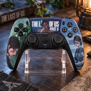 May include: A black wireless video game controller featuring custom graphics from The Last of Us Part II. The centre touchpad displays the game title and a character portrait, while the side grips show detailed illustrations of the same character.