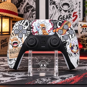 May include: A black and white wireless game controller featuring a custom graphic wrap of One Piece characters, including Monkey D. Luffy in Gear 5 form, alongside wanted poster designs with text reading Dead or Alive and Monkey D. Luffy.
