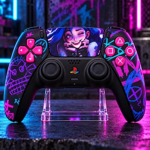 Könnte beinhalten: Ein schwarzer kabelloser Videospiel-Controller mit einer individuellen Grafik eines blauhaarigen Charakters auf dem Touchpad. Das Gehäuse ist mit neonpinken und blauen Graffiti-Kritzeleien, pinken Aktionstasten und einem Logo verziert.
