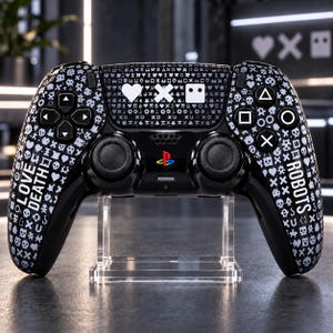 Può includere: Un controller per videogiochi nero con un motivo bianco di piccole icone, tra cui teschi, cuori e croci. Il testo LOVE DEATH è stampato sull'impugnatura sinistra e ROBOTS su quella destra. Un logo PlayStation colorato è posizionato al centro.