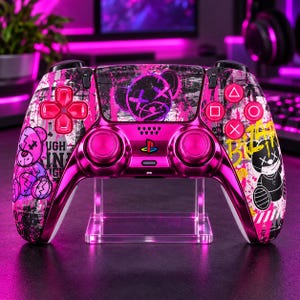 Può includere: Un controller per videogiochi wireless con finitura rosa metallizzato e un design grafico personalizzato. La scocca presenta motivi graffiti in bianco e nero, illustrazioni di orsetti rosa e il testo "TOUGH" e "Bizarre" in grassetto.