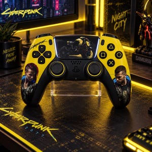 Op de afbeelding: Een gele en zwarte draadloze gamecontroller met Cyberpunk 2077-branding. De controller toont een motorgrafiek op het centrale touchpad, karakterportretten op de handgrepen en het Cyberpunk-logo aan de linkerkant.