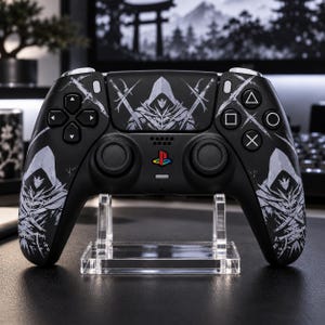 Könnte beinhalten: Ein schwarzer kabelloser Videospiel-Controller mit einem weißen Grafikdruck einer Kapuzenfigur mit gekreuzten Schwertern auf den Griffen und dem zentralen Touchpad. Der Controller verfügt über eine mehrfarbige PlayStation-Logo-Taste.