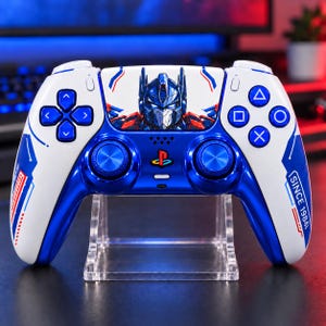Könnte beinhalten: Ein weißer und metallisch blauer kabelloser Gamecontroller mit einer Grafik von Optimus Prime auf dem zentralen Touchpad. Der Controller verfügt über blaue Analogsticks, ein PlayStation-Logo und den Schriftzug SINCE 1984 auf dem rechten Griff.