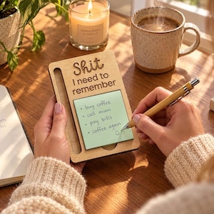 May include: A light-coloured wooden notepad holder with a vertical slot for a pen and a square area for sticky notes. The wood is engraved with the text "Shit I need to remember" above a light green sticky note featuring a handwritten list.