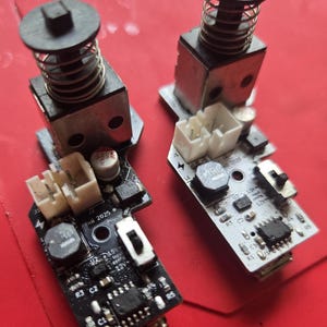 May include: Two electronic circuit boards with black and white bases, each featuring a vertical metal solenoid component with a coiled spring and a black plastic plunger. The boards include white connectors, small capacitors, and printed text labels.