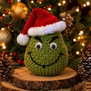 May include: A pear-shaped green crochet plush toy featuring a red and white Santa hat, two plastic googly eyes, and a stitched black curved mouth. The textured green yarn body has a rounded base and a pointed top under the hat.