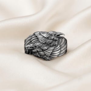 Könnte beinhalten: Ein silberfarbener Metallring mit einem detaillierten, dreidimensionalen Reliefdesign aus zwei Raben mit überlappenden Flügeln und Federn. Die Oberfläche des Rings hat ein dunkles, oxidiertes Finish, das die feinen Texturen der Vögel betont.