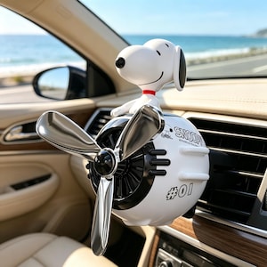Puede incluir: Un ambientador de coche blanco con forma de motor de avión y una figura de Snoopy encima. El dispositivo cuenta con una hélice plateada de tres palas, rejillas de ventilación negras y el texto SNOOPY y #01 impreso en el cuerpo blanco.