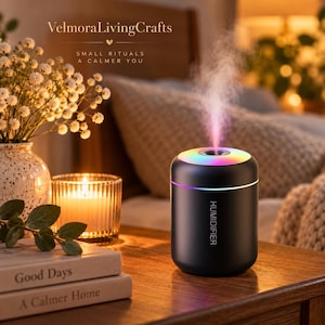 May include: A cylindrical black humidifier with a glowing rainbow-colored LED ring at the top, emitting a fine mist. The device features the word HUMIDIFIER printed in white on the front. It is placed on a wooden surface near books and a candle.