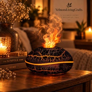 May include: A black, oval-shaped essential oil diffuser with a cracked texture pattern and an orange light strip. It emits a mist illuminated by orange light to resemble a flame. The text VelmoraLivingCrafts A CALMER HOME A BRIGHTER YOU is visible.