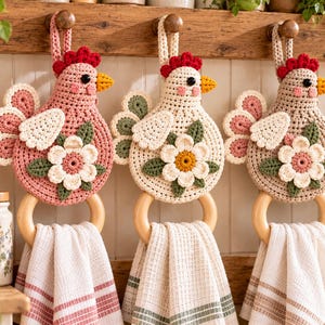 May include: Three crocheted chicken-shaped towel holders in pink, cream, and beige. Each features a red comb, yellow beak, and a white flower appliqué with green leaves. A smooth wooden ring hangs from the base of each chicken, holding a white kitchen towel with colored stripes.