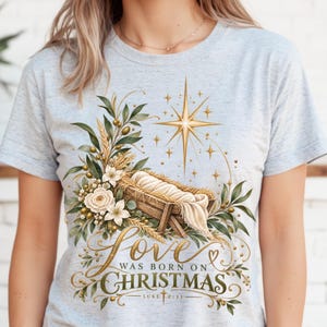 Op de afbeelding: Een lichtgrijs gemêleerd T-shirt met een afbeelding van een kribbe met een ingebakerde baby, omringd door groen, witte bloemen en een gouden ster. De tekst luidt: Love was born on Christmas Luke 2:11 in elegante gouden letters.