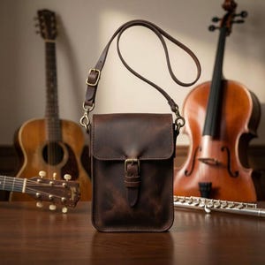 Pode incluir: Uma bolsa tiracolo de couro castanho escuro com formato retangular vertical, aba dobrável e uma alça ajustável. A bolsa apresenta um fecho de fivela central feito de ferragens de metal em tom de latão antigo e costuras visíveis.