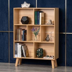 May include: A light wood bookshelf with six open compartments, supported by four tapered legs. The shelves contain books, a black geometric object, a crystal cluster, a glass vase with lavender, a bird figurine, and a decorative bowl.