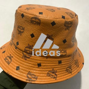 Puede incluir: Un sombrero de pescador color canela con superficie texturizada que presenta un patrón repetitivo de diamantes y el logotipo MCM en negro. El frente muestra un gráfico gris de tres barras inclinadas sobre el texto blanco en minúsculas ideas.