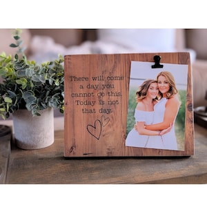 May include: A rustic wooden picture frame with a black clip holding a photo of two women hugging. The frame has the text "There will come a day you cannot do this. Today is not that day." engraved on it.