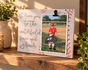 May include: A rectangular wooden photo frame with a light background featuring red baseball stitching patterns on the sides. The text reads, I love you to the centerfield fence and back, with a heart icon. A black clip holds a photo of a baseball player.