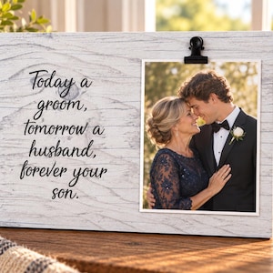 May include: A rectangular white wood-grain photo frame featuring a black metal clip holding a photograph of a person in a dark blue lace dress embracing a person in a black tuxedo. The frame displays the black text: Today a groom, tomorrow a husband, forever your son.