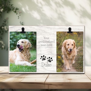 Op de afbeelding: Wit houten fotolijst met twee openingen voor foto's. De lijst heeft een clip bovenaan elke opening. De lijst heeft een pootprintontwerp en de tekst "Your personalized text here" en "Duke". De lijst staat op een houten oppervlak. Twee golden retriever honden staan op de foto's.
