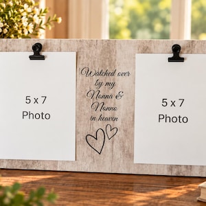 May include: A rectangular light wood-grain photo frame featuring two black metal clips for holding 5x7 inch photos. The center panel displays the black text "Watched over by my Nonna & Nonno in heaven" above two hand-drawn heart outlines.