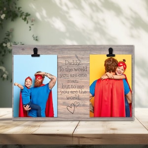 May include: A wooden picture frame with two photo slots, each held by a black clip. The frame features a photo of a father and child dressed as superheroes. Text on the frame reads "Daddy. To the world you are one man, but to me you are the world."