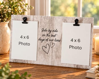 Op de afbeelding: Een rechthoekige fotolijst met een lichte houtnerf en twee zwarte metalen clips die witte papieren inzetstukken met het label 4 x 6 Photo vasthouden. Het midden bevat zwarte tekst Side by side on the best days of our lives en twee hartjes.