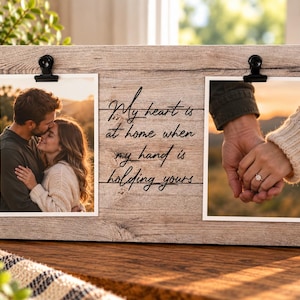 May include: A rectangular wooden photo frame with a light brown wood grain finish, featuring two black metal clips to hold photographs. The center of the frame displays the black text: My heart is at home when my hand is holding yours.