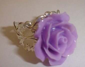 Peut inclure: Bague argentée ornée d'une grande rose lavande. La rose est le point central, avec des pétales détaillés. La monture de la bague présente un motif filigrané orné. La bague est posée sur un fond neutre.