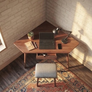 May include: A wooden corner desk with a trapezoidal top, a single central drawer, and four tapered legs. The desk holds a laptop, a black desk lamp, a small potted succulent, a spiral-bound notebook, a pen, and a smartphone. A grey stool sits in front.