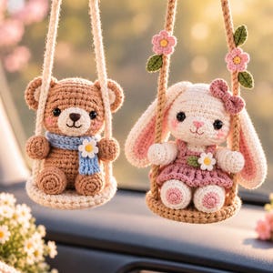 May include: Two crocheted hanging ornaments featuring a brown bear and a white rabbit sitting on small swings. The bear wears a blue scarf with a white flower, while the rabbit wears a pink dress with a white flower and a pink bow on its ear.