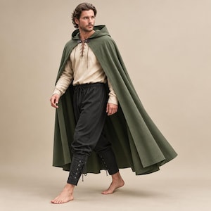 Könnte beinhalten: Ein bodenlanger, olivgrüner Kapuzenumhang über einem beigen Langarmshirt mit Schnürung, einer schwarzen Hose und schwarzen, geschnürten Wadenwickeln. Der Umhang ist am Hals mit einem dunkelbraunen, lederartigen Verschluss versehen.